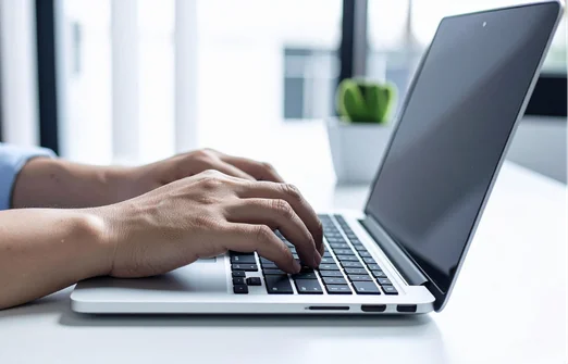 Close-up of hands typing on a laptop keyboard on a white desk with a blurred plant in the background.