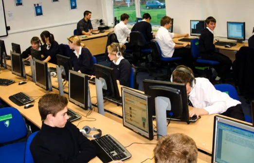Students in school uniforms working on desktop computers in a classroom with several rows of desks.