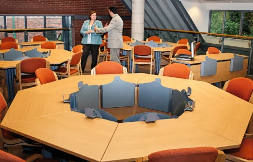 Two professionals talking in a modern office space with octagonal wooden desks and orange chairs.