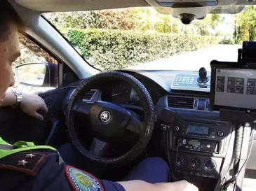 Police officer sitting in the driver's seat of a patrol car with dashboard and electronic devices visible.