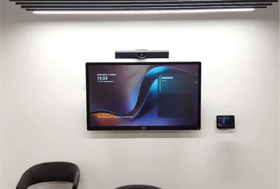Wall-mounted large screen showing meeting availability with two black chairs below it in a plain room.