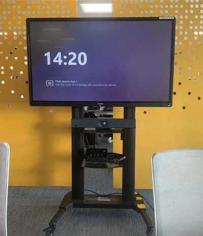 Large TV on wheeled stand showing 14:20 and meeting room instructions in office space.