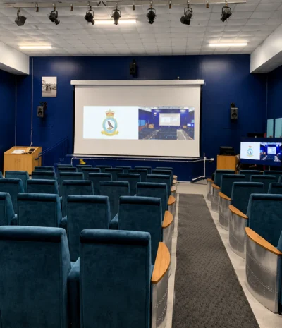 Empty auditorium with blue chairs facing a screen displaying a logo and a stage area.
