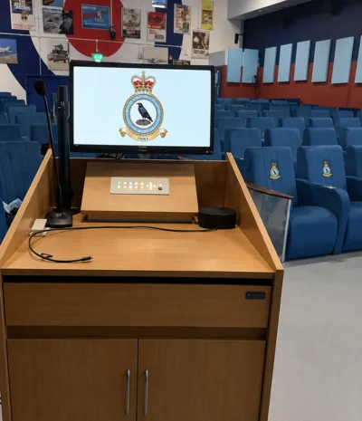 Podium with microphone and screen showing RAF emblem in an empty auditorium with blue seats.