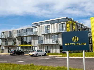Modern three-story building with large windows, black and yellow signage reading BATTLELAB, and parked cars outside.