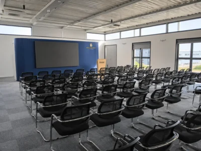 Empty modern classroom with rows of black chairs facing a podium and large screen on the front wall.