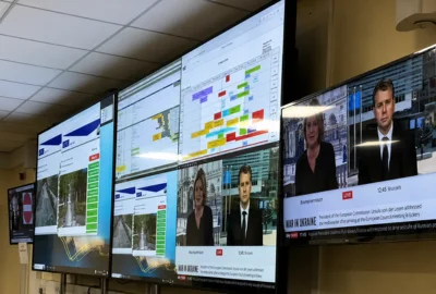 Multiple screens on a wall showing news broadcasts and data charts in a control room.