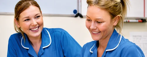 Two female healthcare workers in blue uniforms smiling and engaging in conversation.