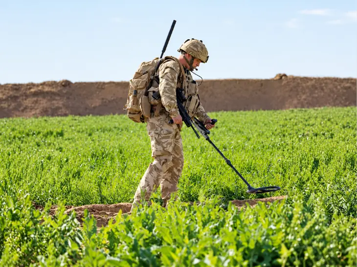 Soldier in camouflage using a metal detector to scan soil in a green field.