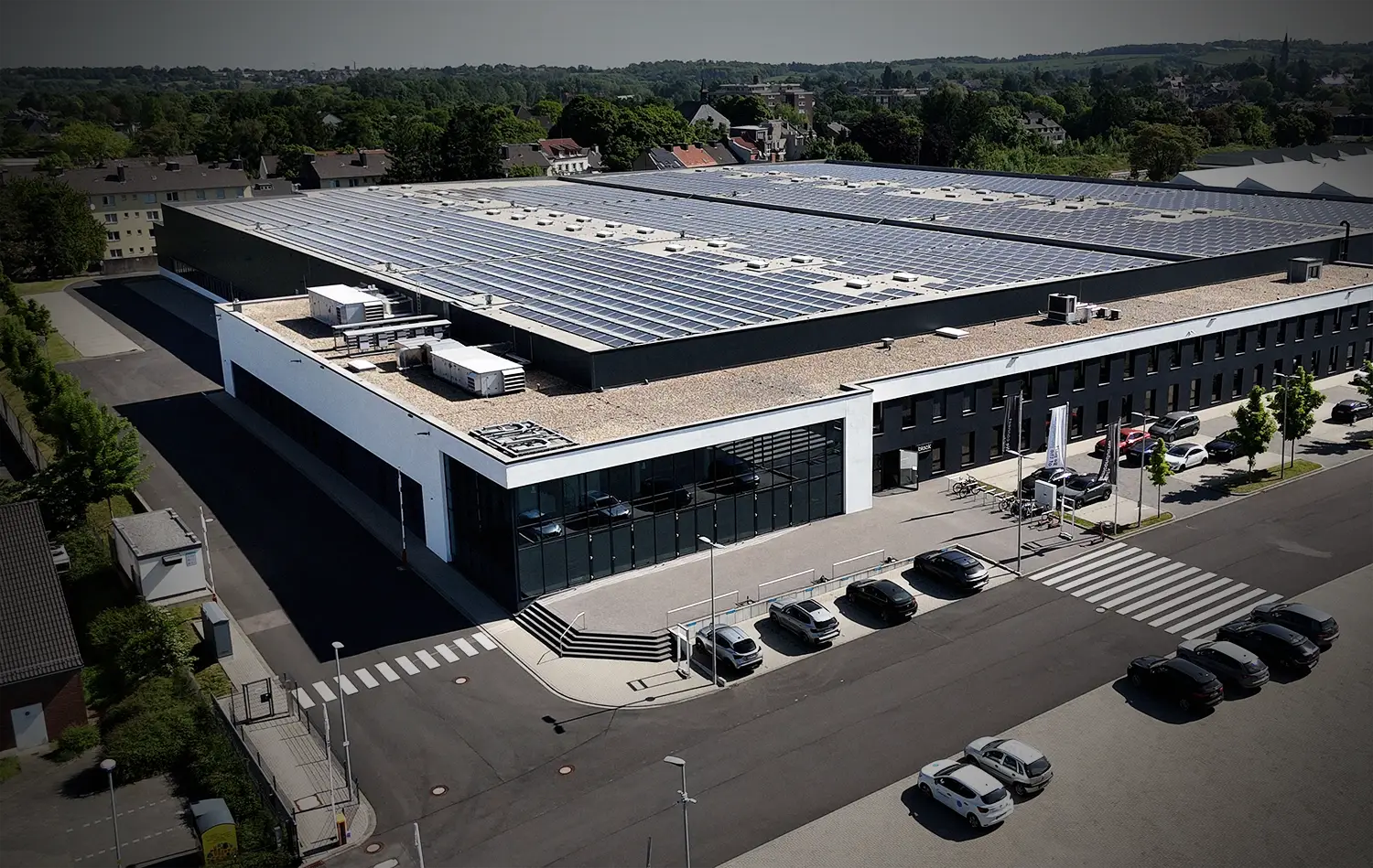 Aerial view of a large industrial building with solar panels on the roof and cars parked in front along the street.
