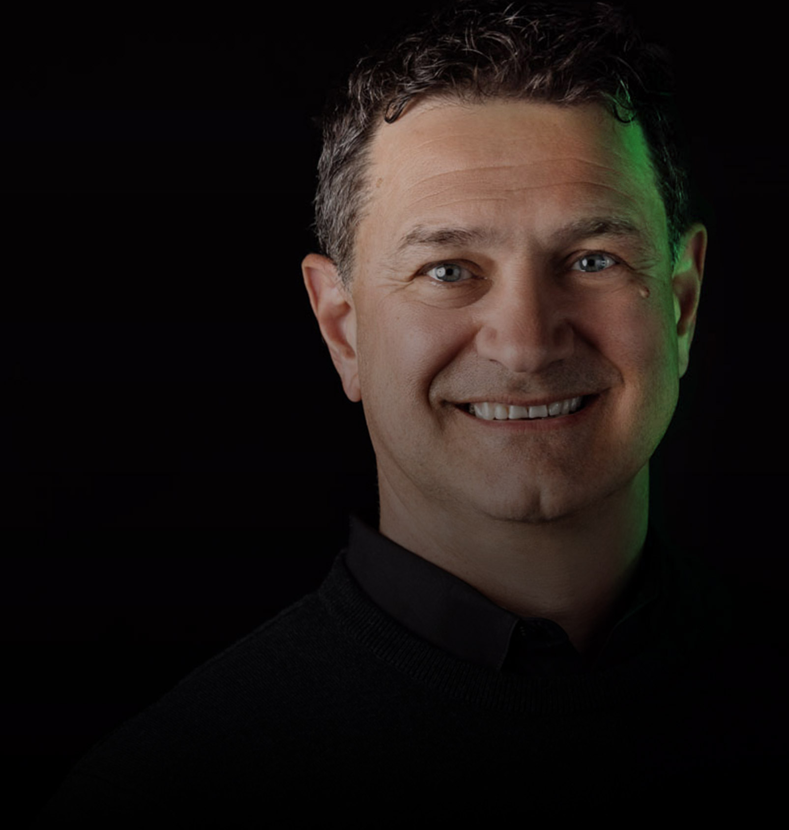 Smiling man with curly dark hair wearing a black sweater, against a dark background with a subtle green light on the right side of his face.