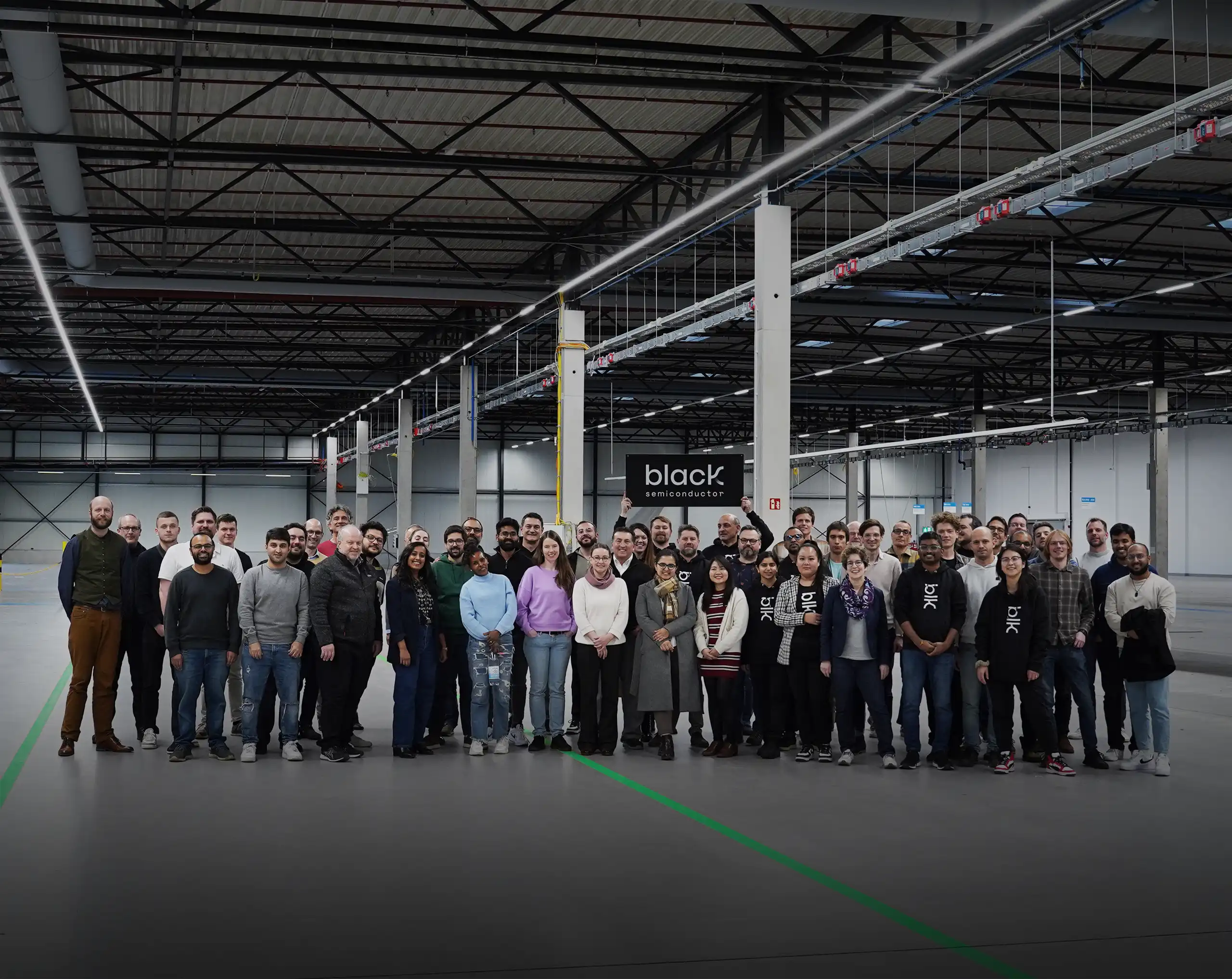 Group of diverse people standing in a large industrial warehouse holding a 'black semiconductor' sign.