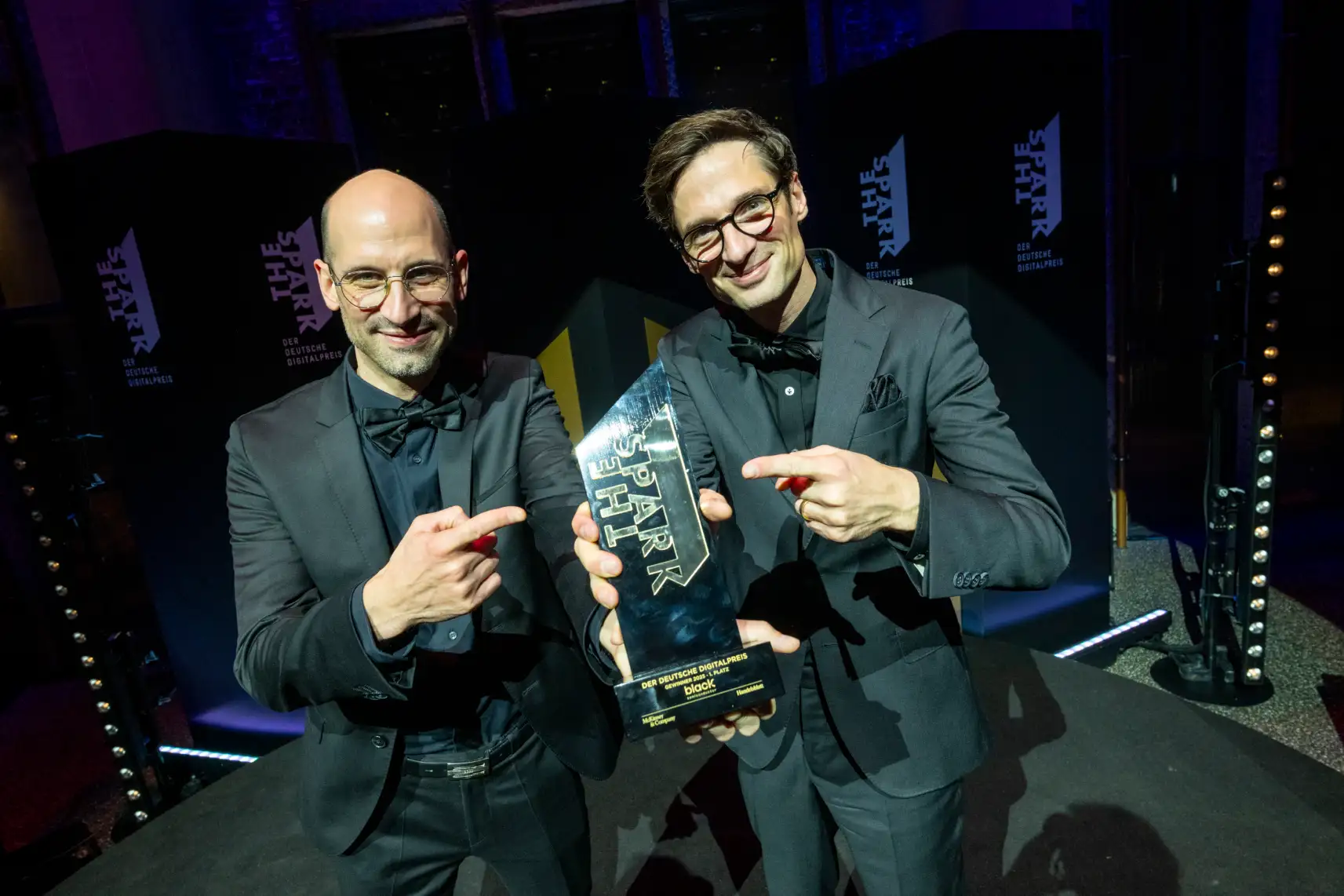 Two men in black suits and bow ties smiling and pointing at a trophy labeled 'The Spark' at an award ceremony.