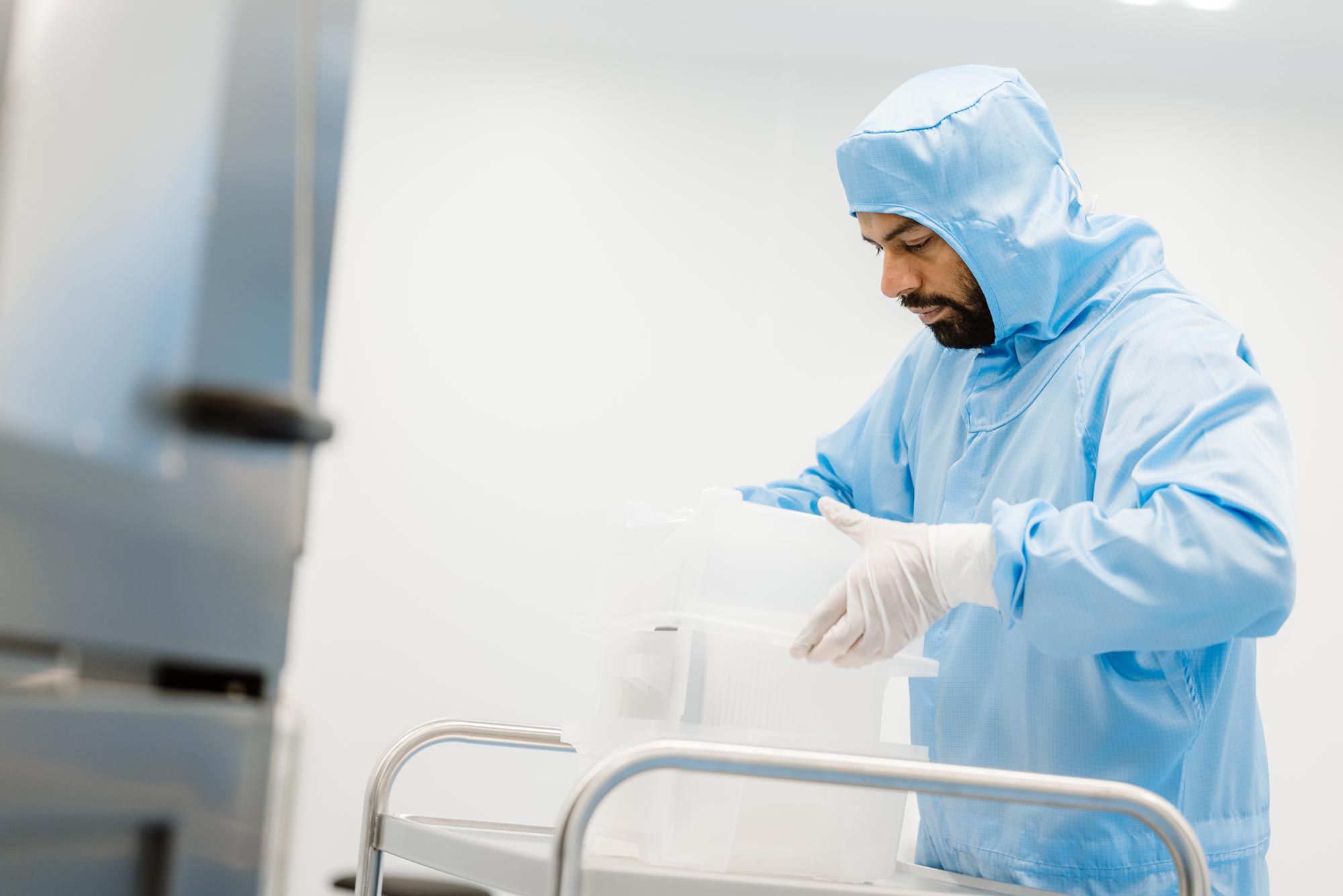 Technician in blue cleanroom suit and gloves handling plastic containers in a semiconductor lab.