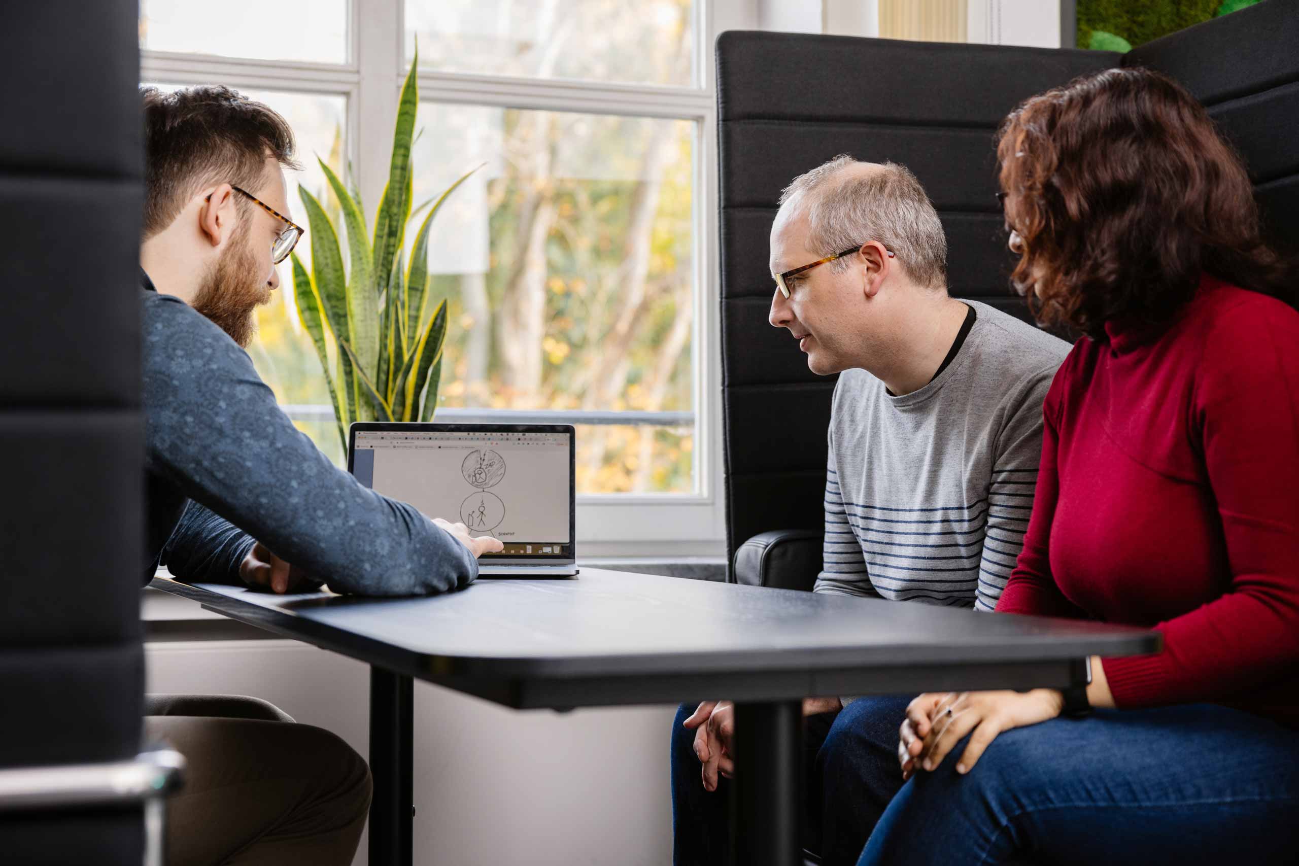 Three colleagues sitting at a table in an office, looking at a laptop screen with sketches.