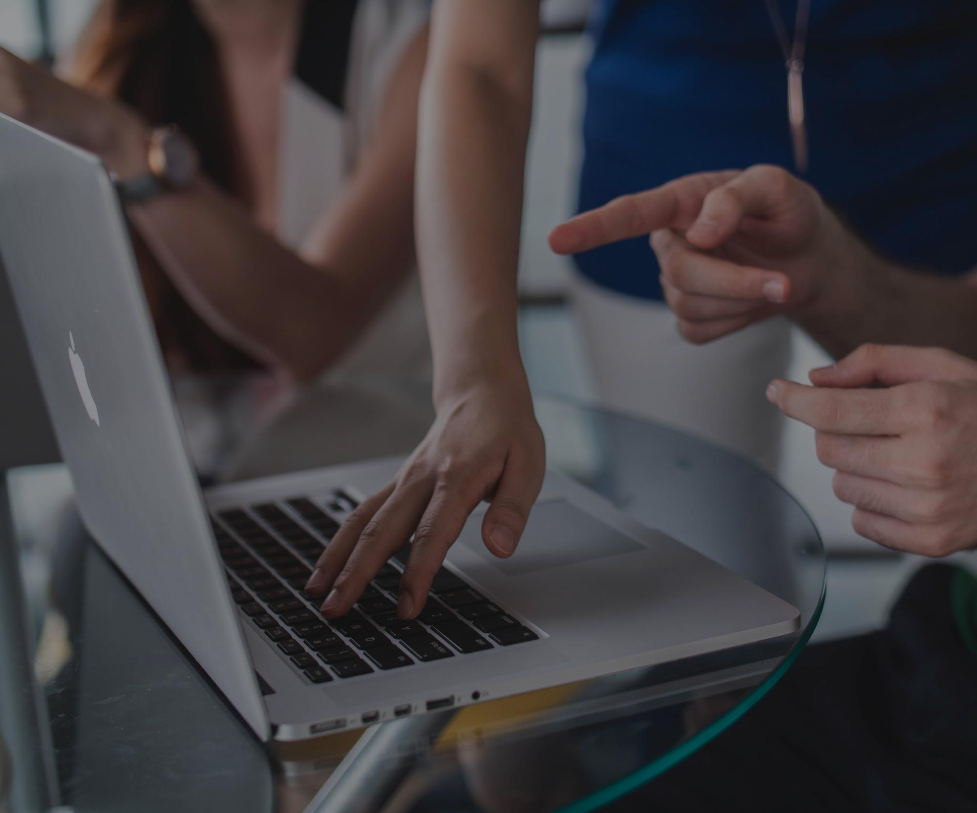 Two people working together on a silver MacBook laptop on a glass table, one typing and the other pointing at the screen.