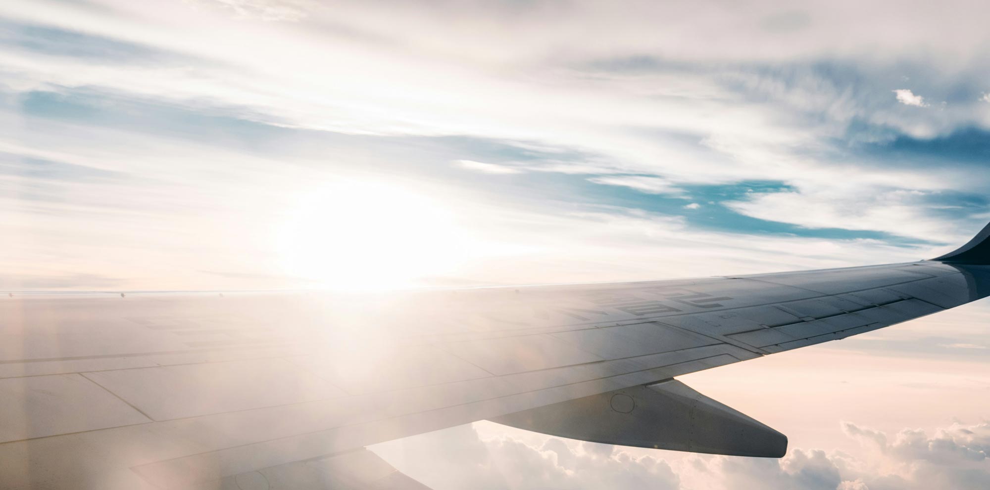 Airplane wing with the sun shining brightly and clouds below during flight.