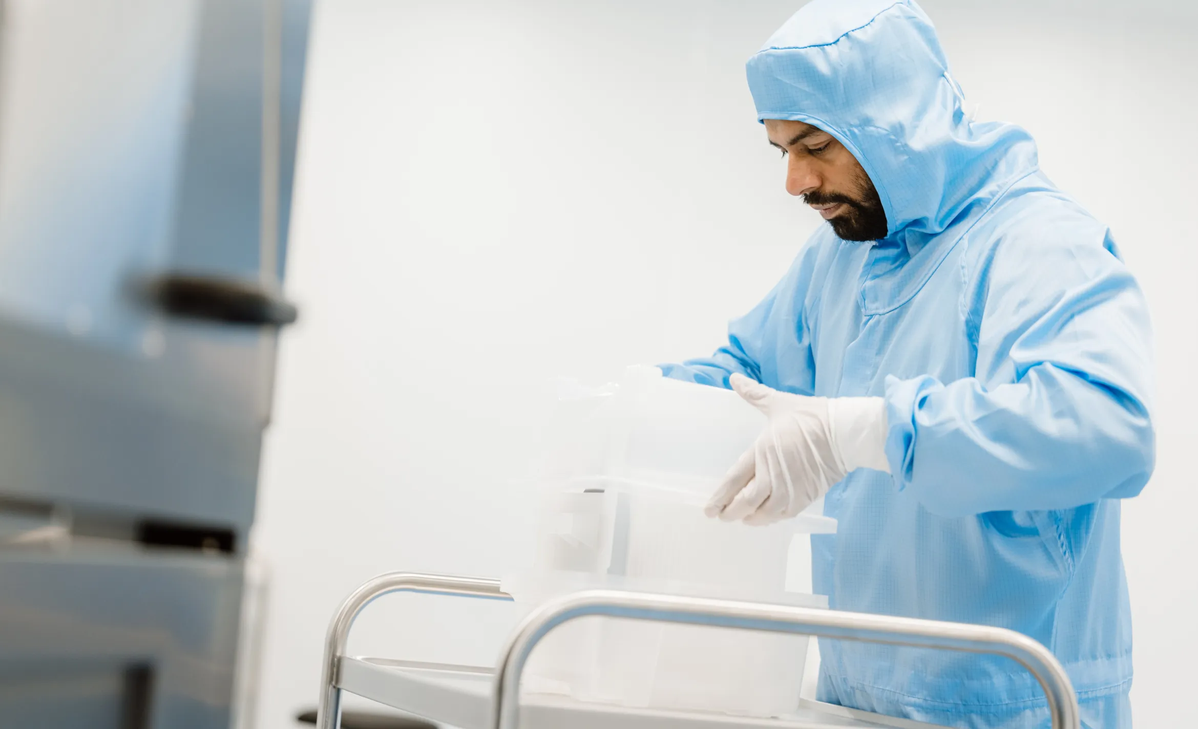 A man wearing a blue cleanroom suit and white gloves working with translucent plastic containers on a metal cart in a sterile laboratory environment.