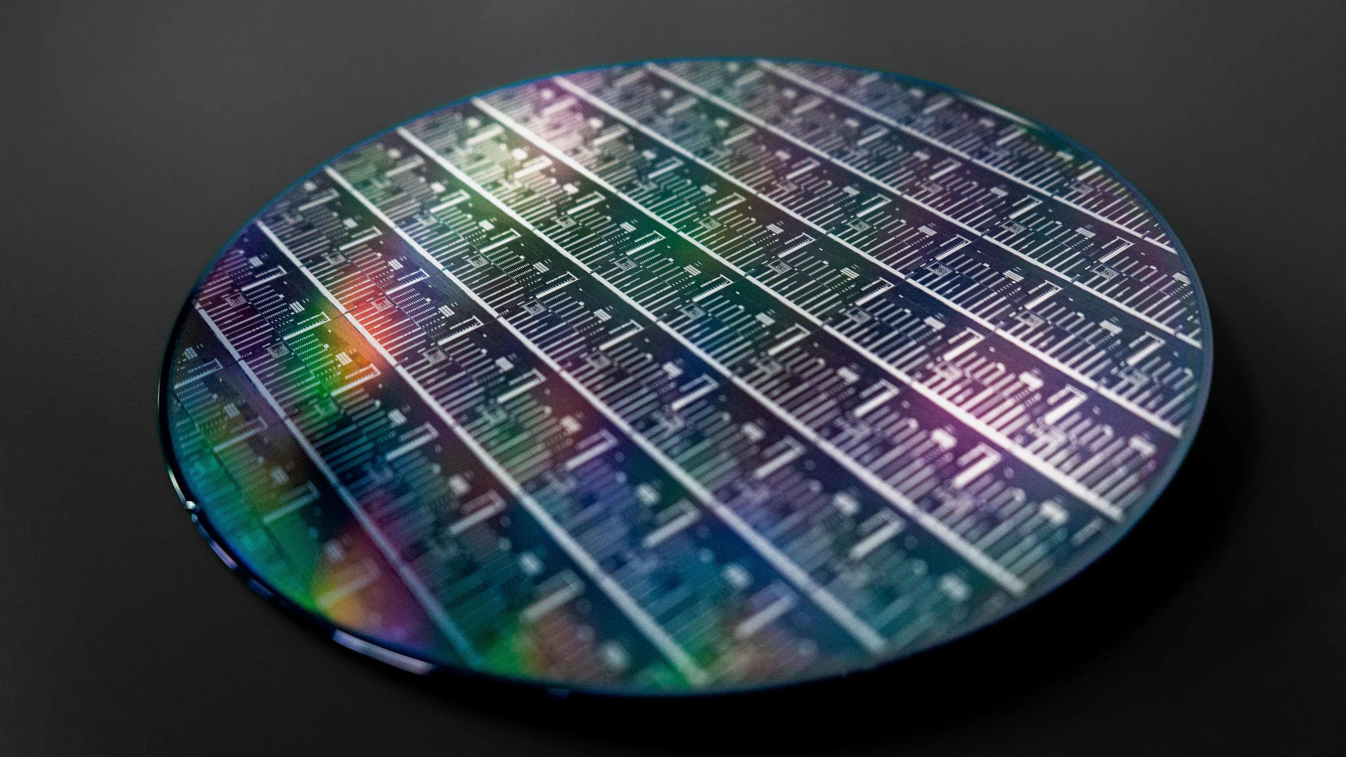 Close-up of a circular silicon wafer with multiple rectangular microchip patterns, showing a reflective surface with subtle rainbow hues against a dark background.