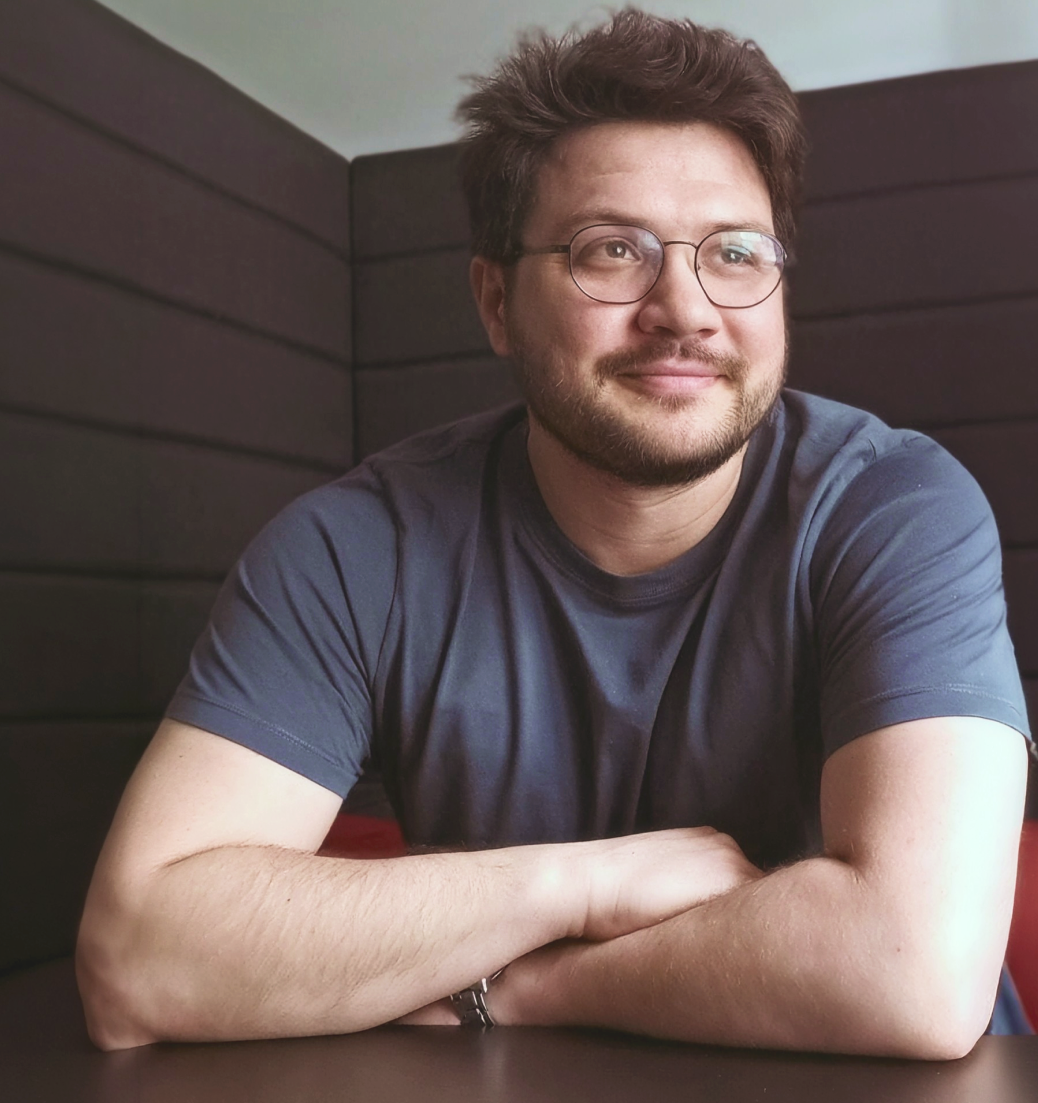 A man with glasses, short dark hair, and beard wearing a dark gray t-shirt, smiling softly while sitting with his arms resting on a surface, against a background of black padded walls.