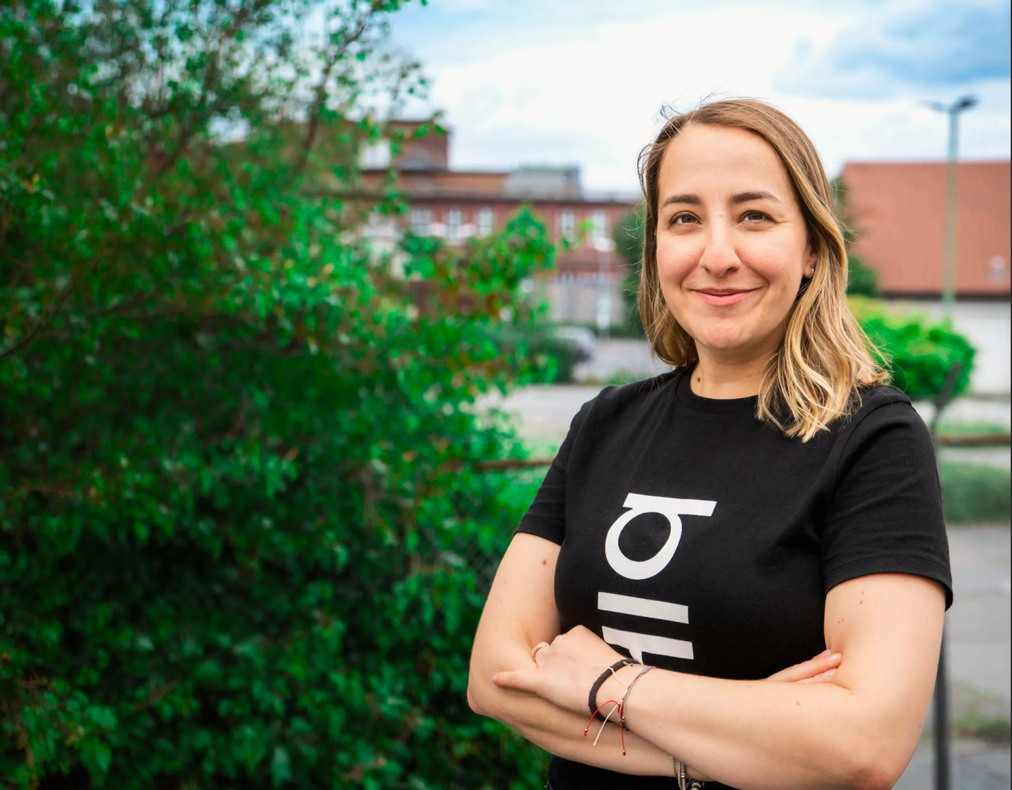 A smiling woman with blonde hair wearing a black t-shirt with white text, standing outdoors with green bushes and buildings in the background.
