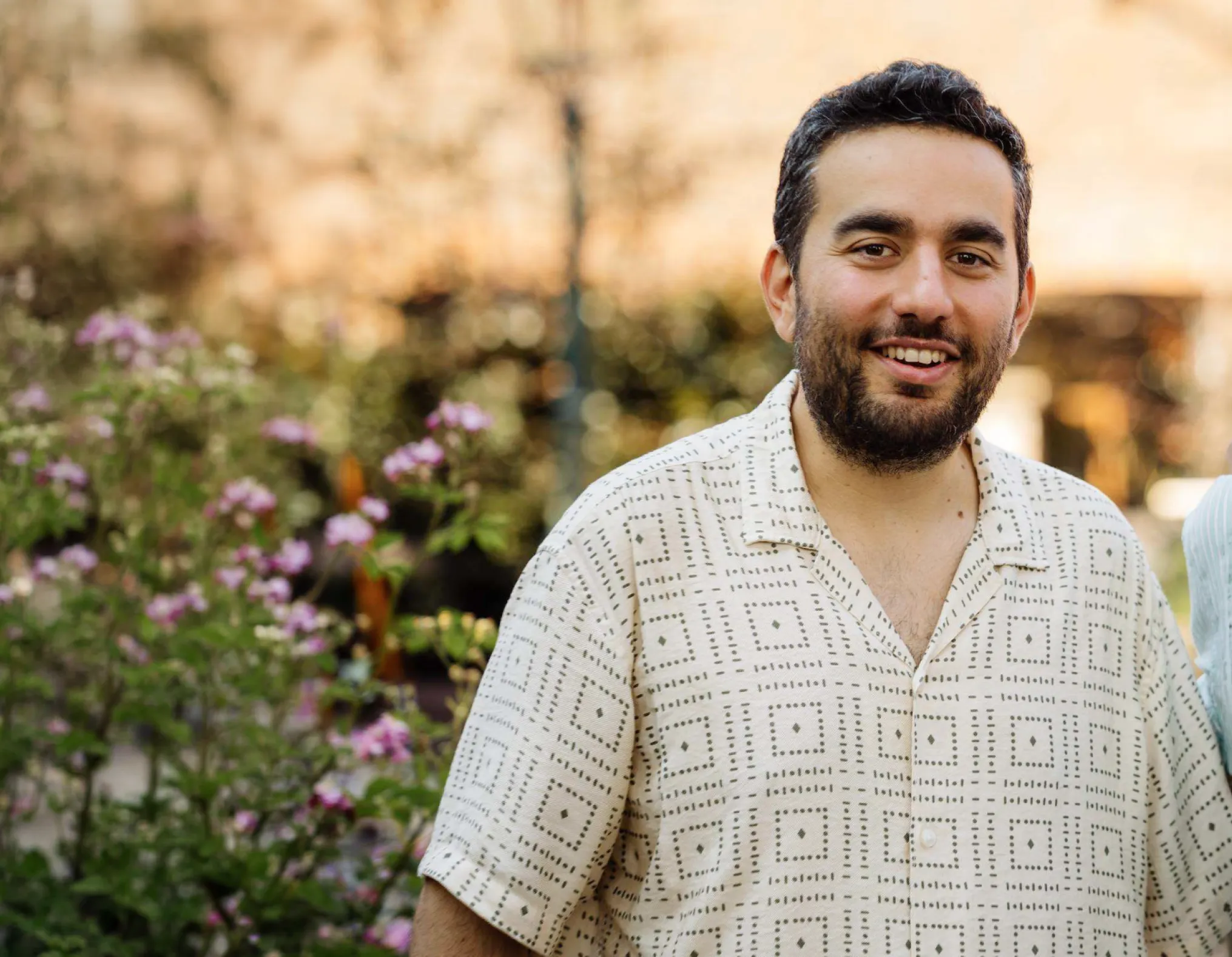 Smiling man with short curly dark hair and beard wearing a beige patterned short-sleeve shirt stands outdoors with green plants and pink flowers blurred in the background.