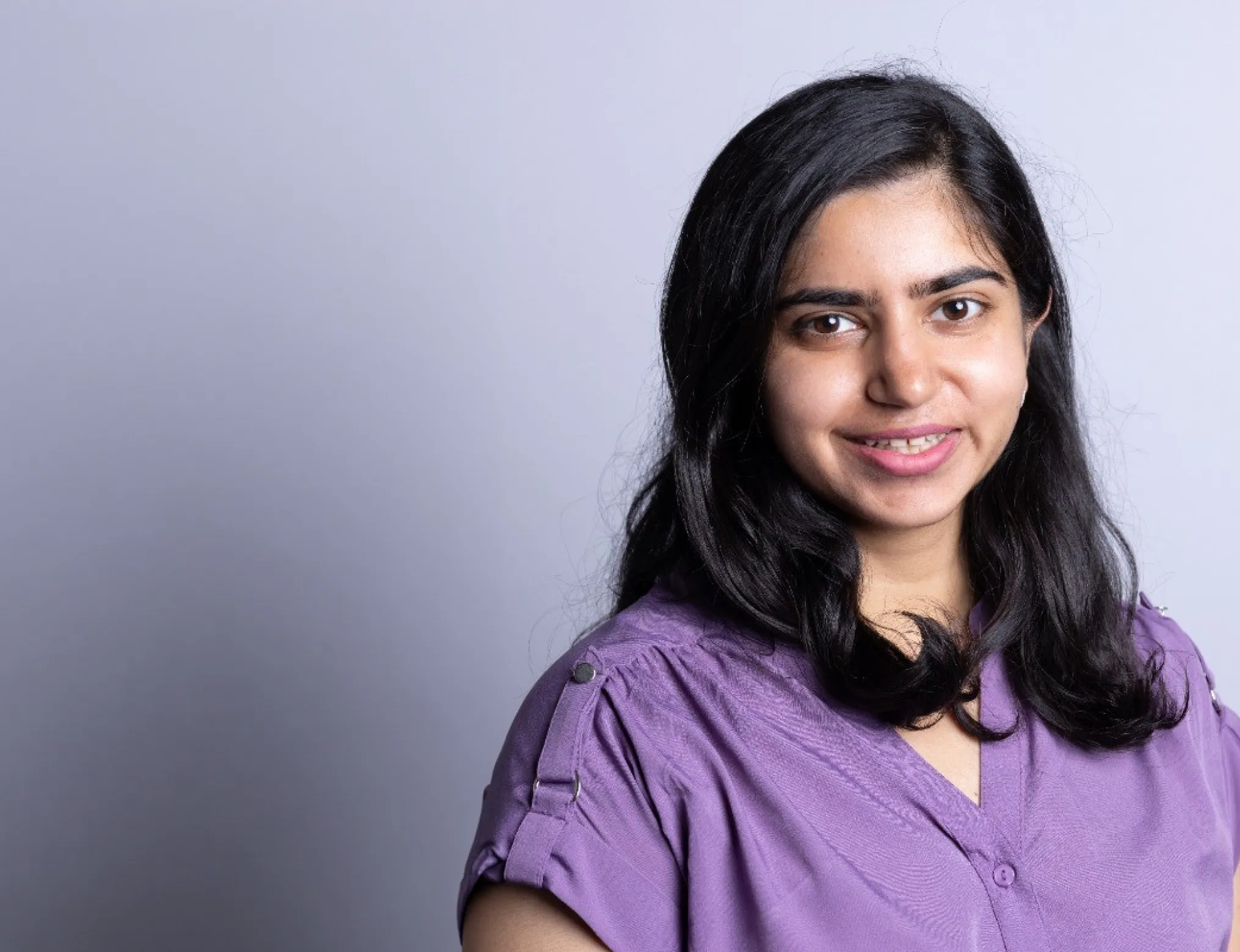 Woman with medium-length black hair wearing a purple blouse smiling against a plain light gray background.