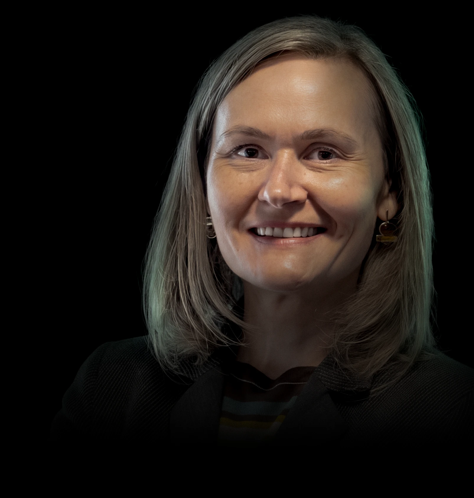 Smiling woman with shoulder-length blonde hair wearing a black blazer and dangling geometric earrings against a black background.