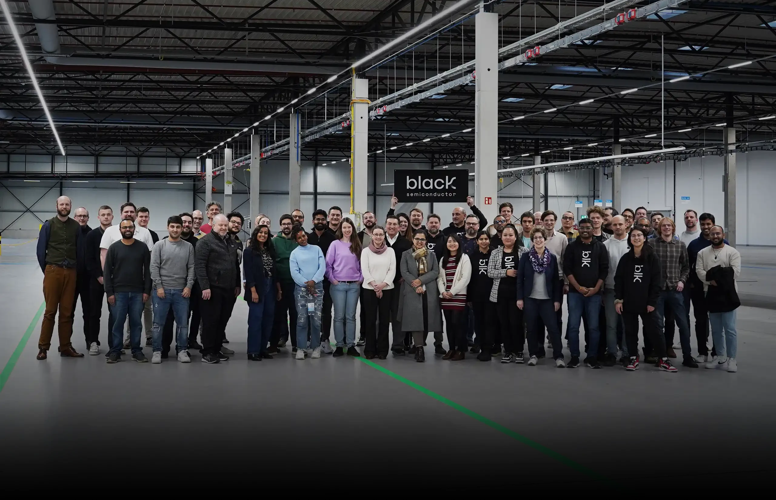 Large group of people standing together in a spacious, industrial warehouse setting, with one person holding a black sign that reads 'black semiconductor'.