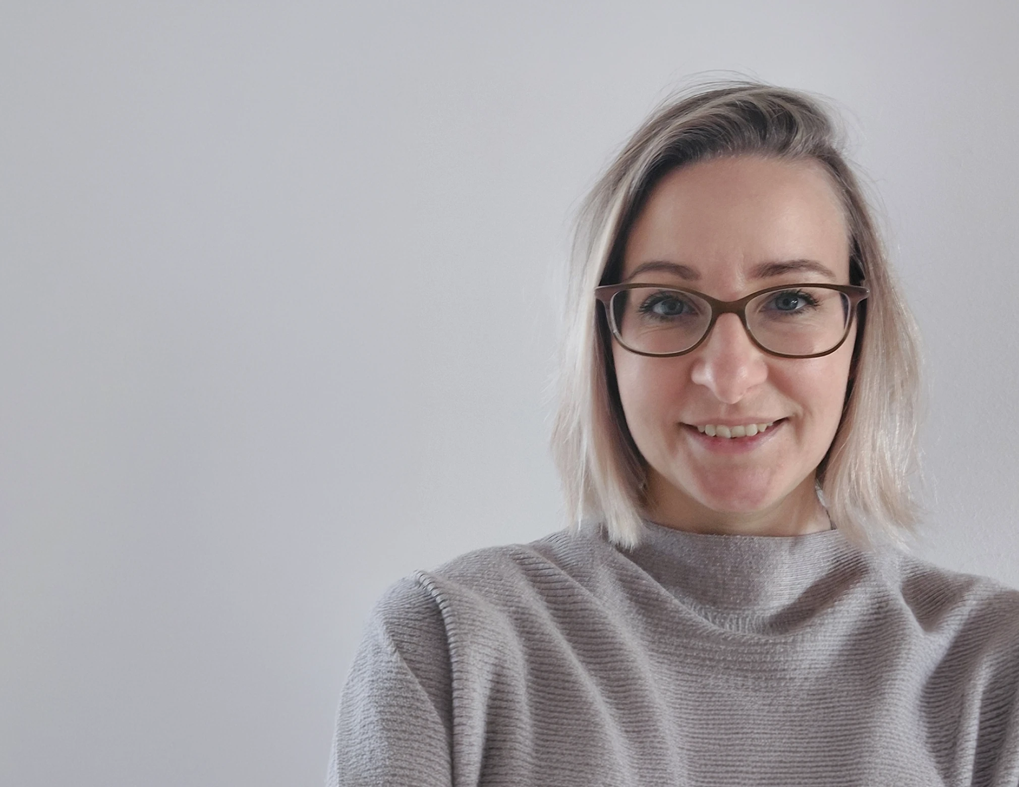 Smiling woman with shoulder-length blonde hair wearing brown glasses and a light gray sweater standing against a plain light gray background.
