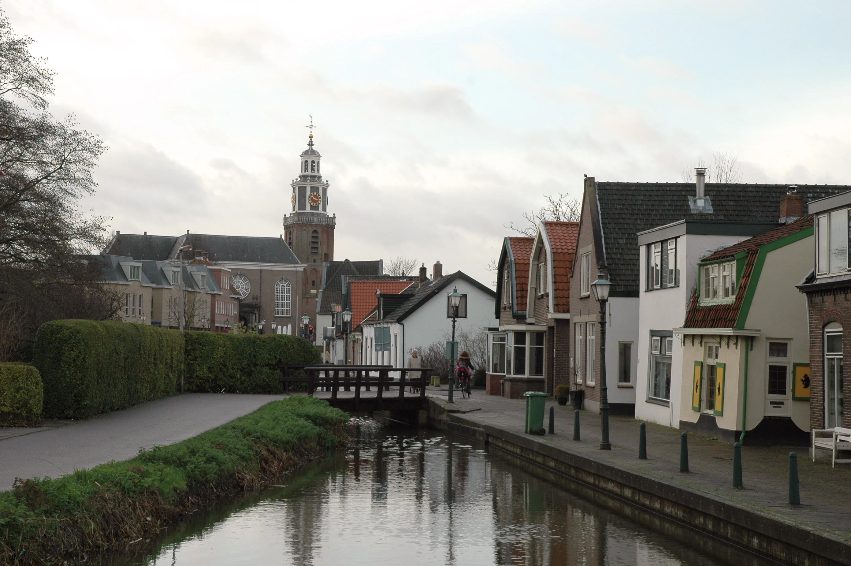 Quiet canal in a small town bordered by green bushes and a sidewalk with traditional Dutch houses on the right, a wooden footbridge crossing the canal, and a tall church tower with a clock in the background under a cloudy sky.