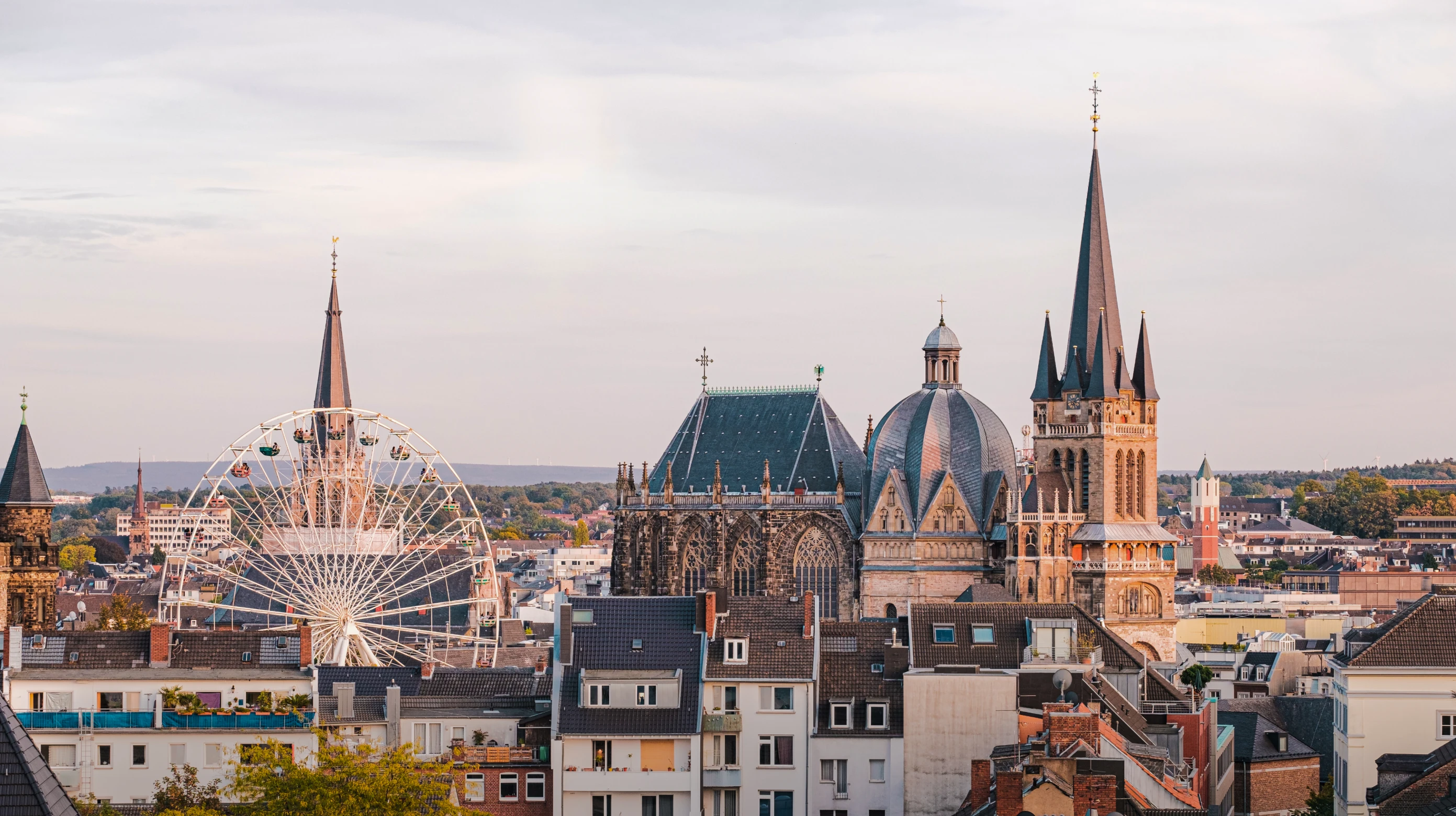 Cityscape of Aachen, Germany, featuring historic buildings with gothic architecture, pointed church spires, a large ferris wheel, and residential rooftops under a cloudy sky.