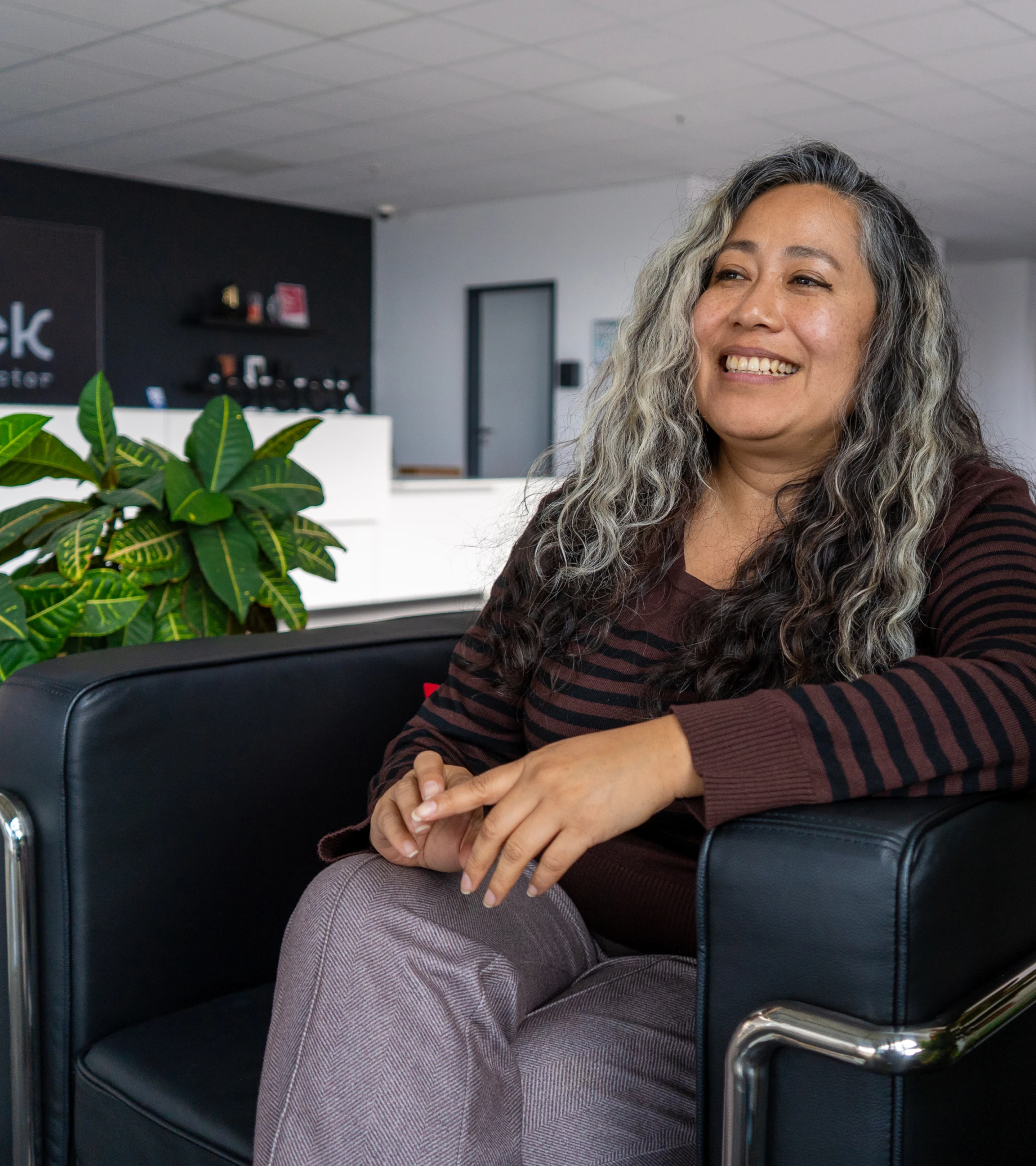 Smiling woman with long, wavy gray and black hair sitting in a black armchair in a modern office with a large green plant and a sign reading 'black semiconductor' in the background.