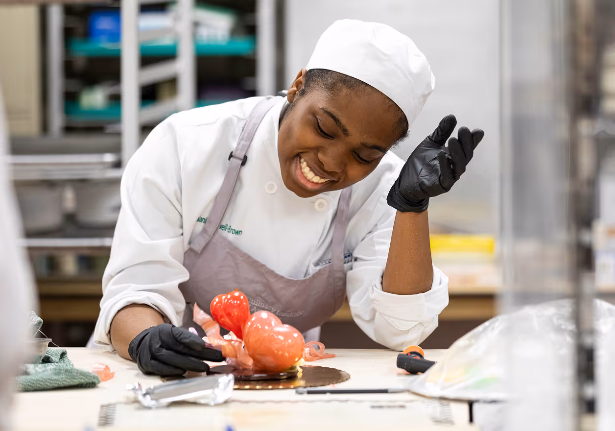 Smiling chef wearing a white hat and apron sculpting a red sugar art piece on a worktable.