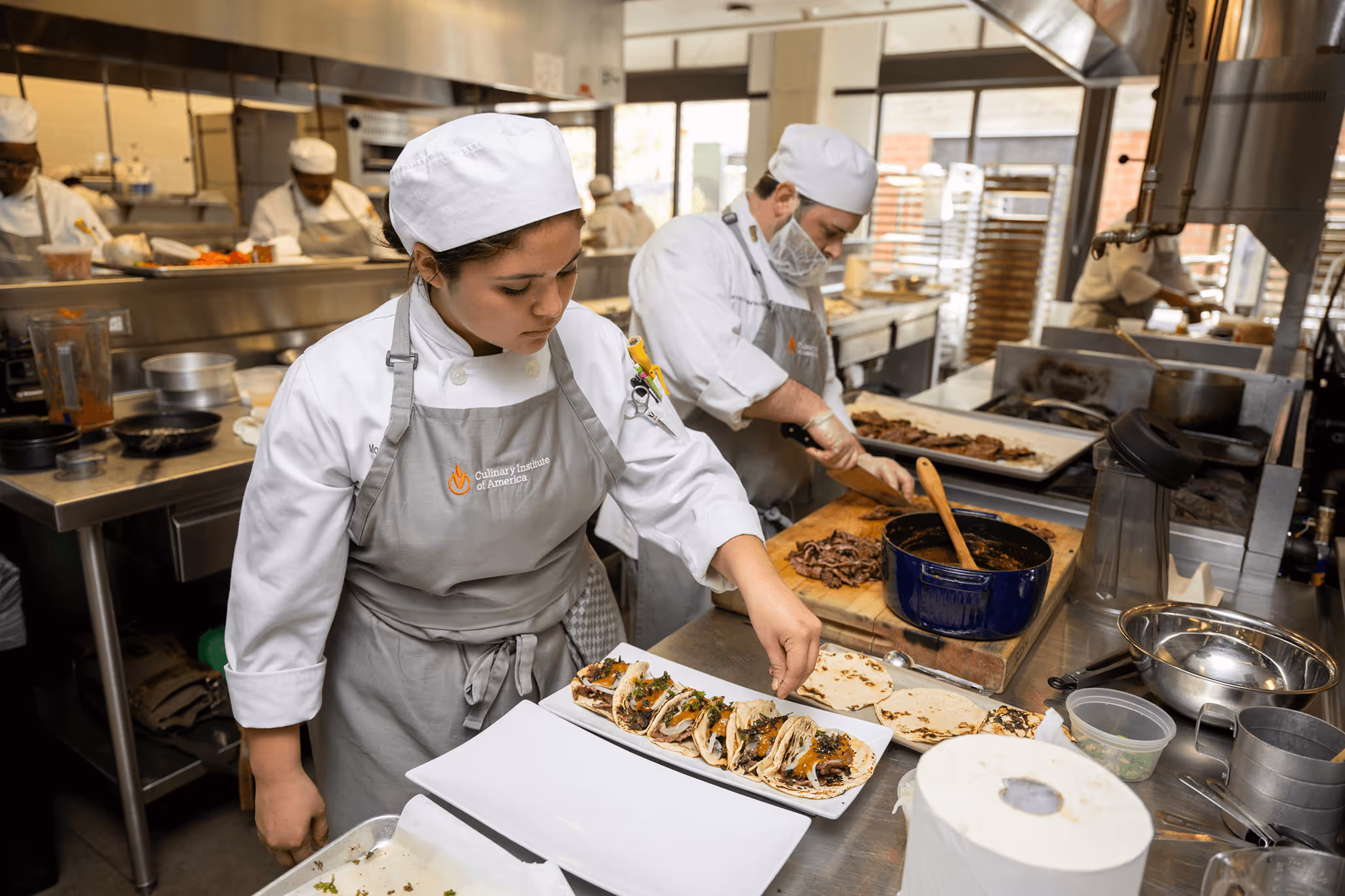 Two chefs in Culinary Institute of America uniforms prepare tacos and slice meat in a commercial kitchen.