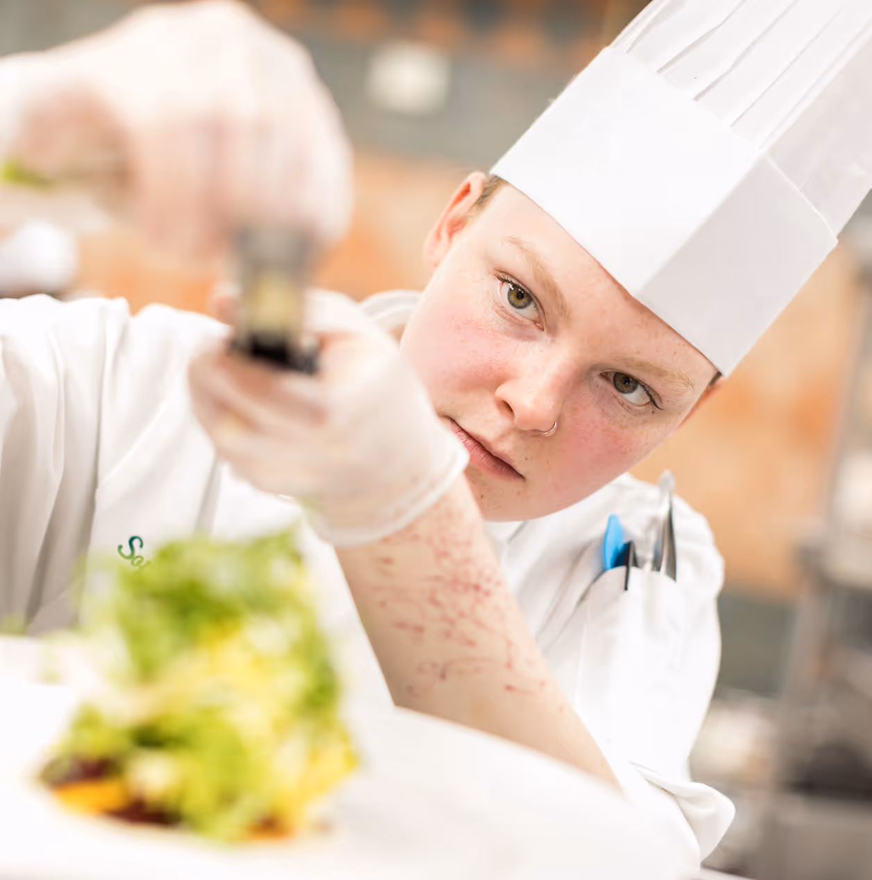Chef wearing a white hat and uniform intensely focusing while seasoning a dish with a pepper grinder in a kitchen.
