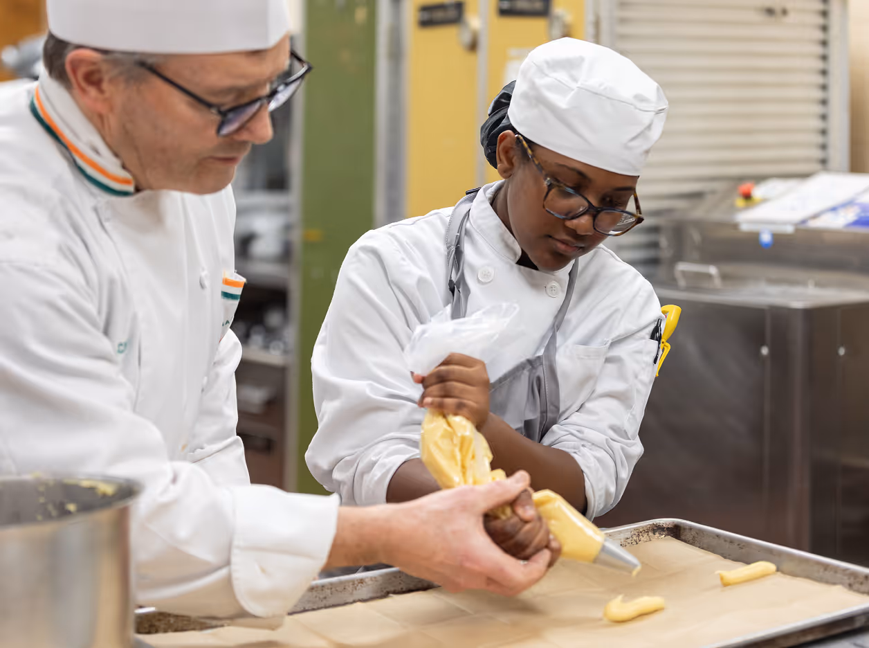 Two chefs in white uniforms and hats piping dough onto a baking sheet in a professional kitchen.