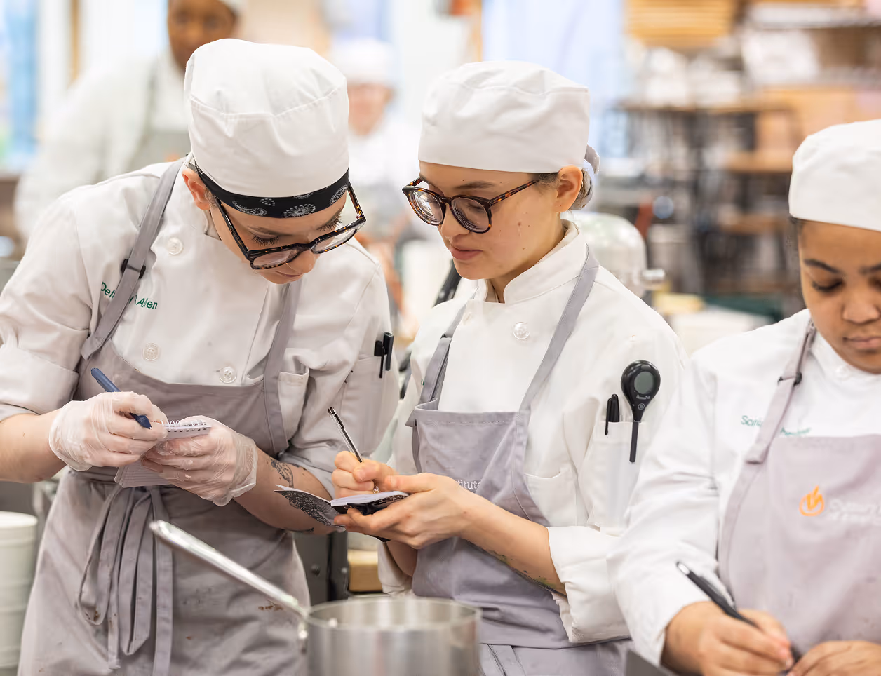 Three chefs wearing white uniforms and aprons writing notes in a busy kitchen.
