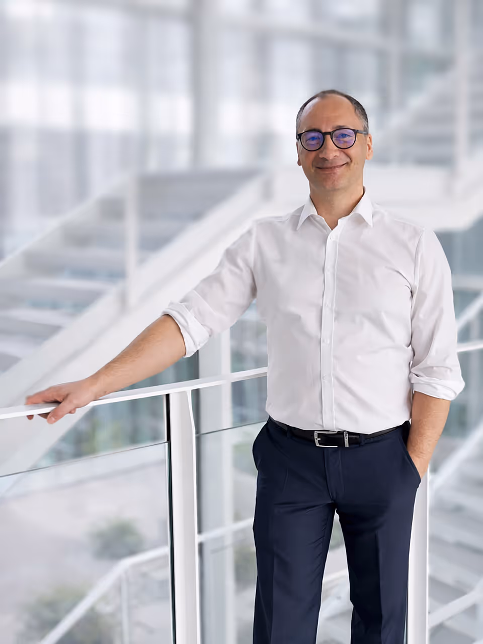 Ein freundlich lächelnder Herr Sascha Korthals mit weißem Hemd und dunkler Hose lehnt sich an einem Geländer vor einem unscharfem modernen Innenraum.