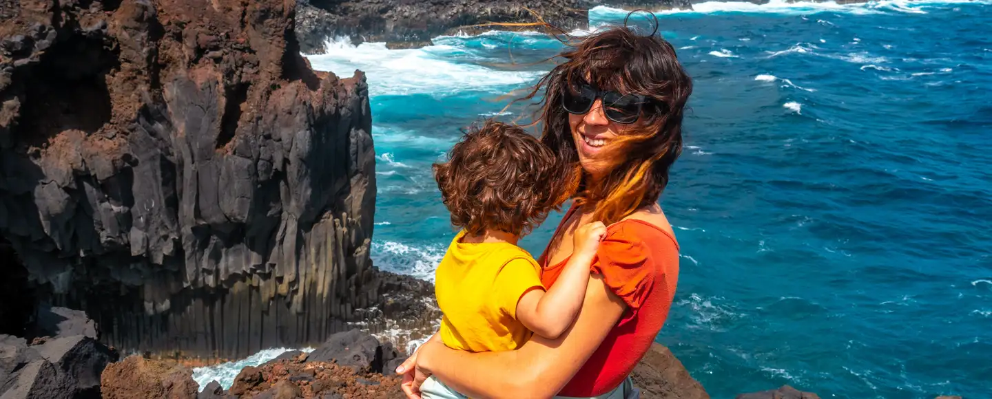 une femme porte un enfant dans ses bras devant un paysage de mer avec des falaises, elle est partie en vacances en octobre
