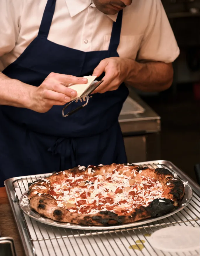 Person in white shirt and dark apron grating cheese over a freshly baked pizza with a charred crust.