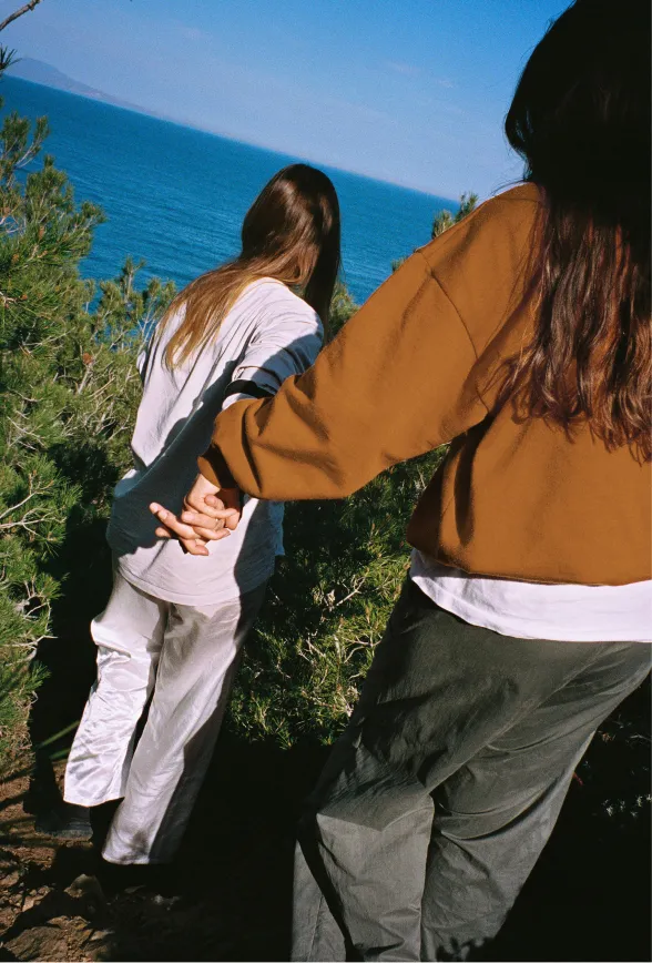 Two people holding hands walking along a greenery area near the sea under a clear blue sky.