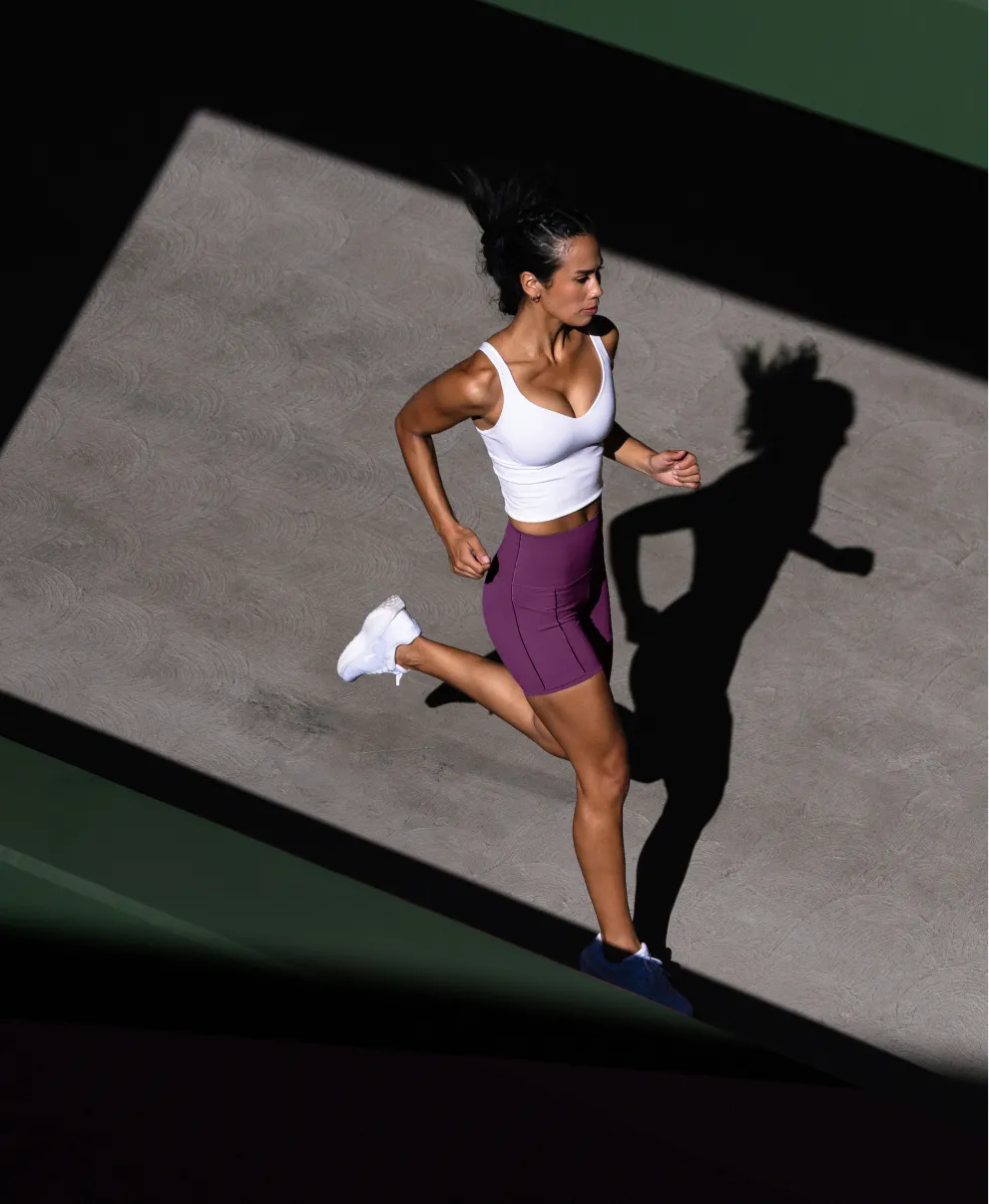 Woman running outdoors wearing a white tank top, purple shorts, and white sneakers casting a shadow on the ground.
