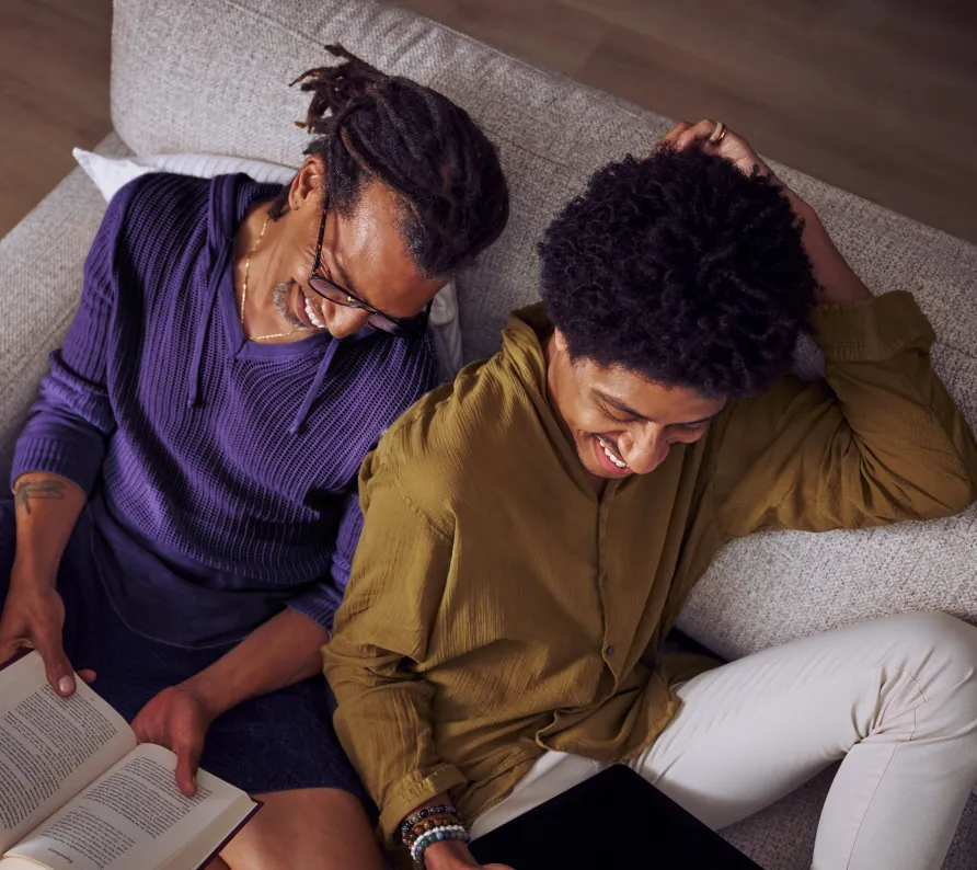 Two people sitting close on a couch, one reading a book and both smiling warmly.