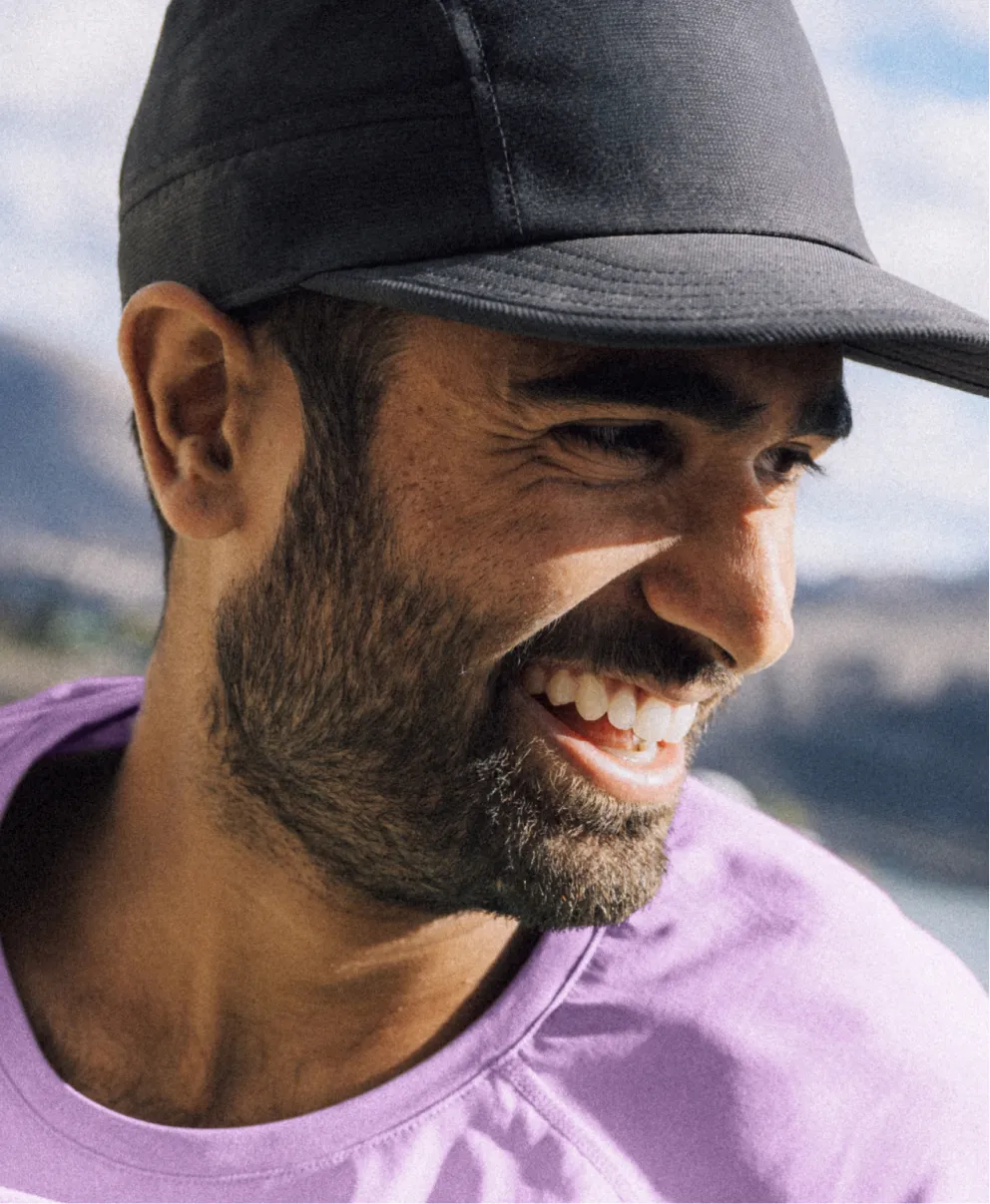 Bearded man wearing a dark cap and light-colored shirt smiling outdoors.