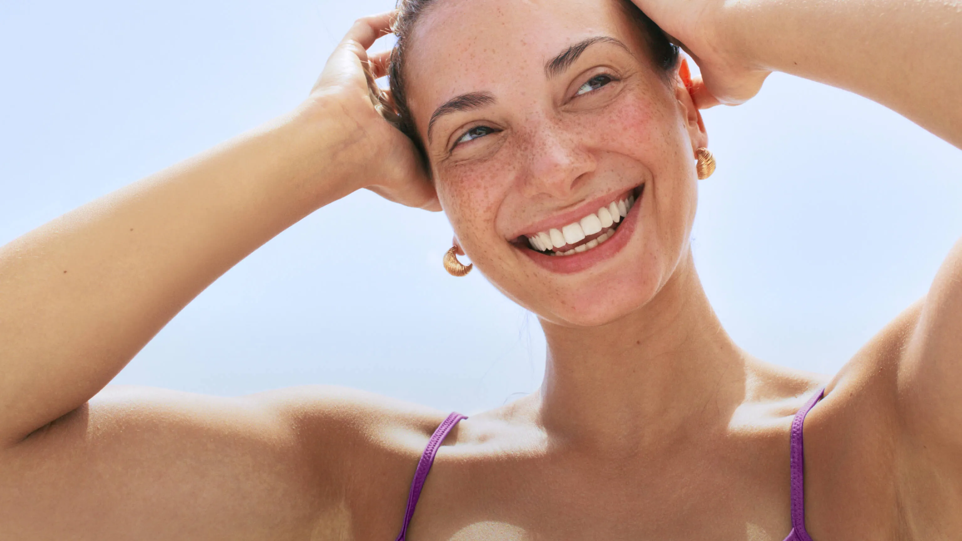 Smiling woman with earrings and purple straps holding her hair back against a clear blue sky.