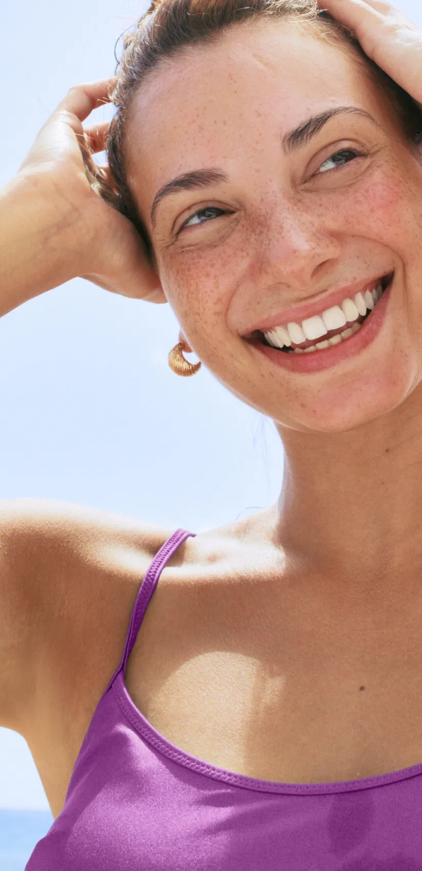 Smiling woman with earrings and purple straps holding her hair back against a clear blue sky.