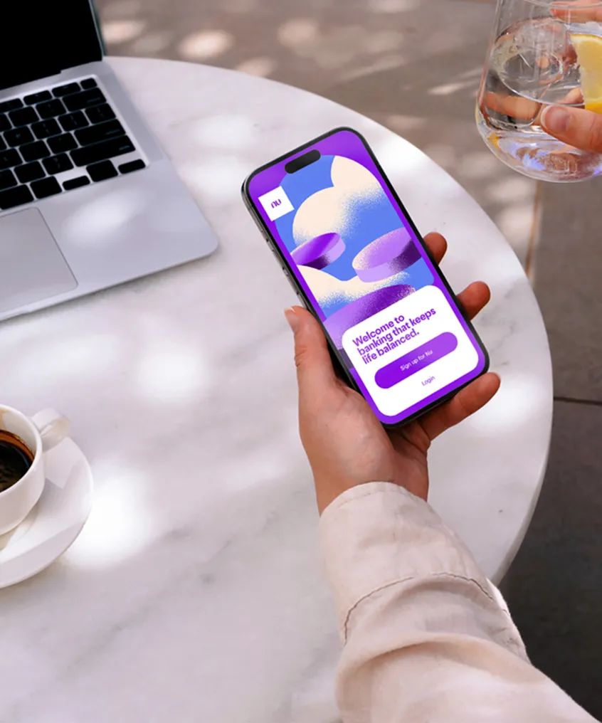 Person holding a smartphone displaying a package tracking app with a hot air balloon graphic, near a glass of water with lemon and a laptop on a round table.