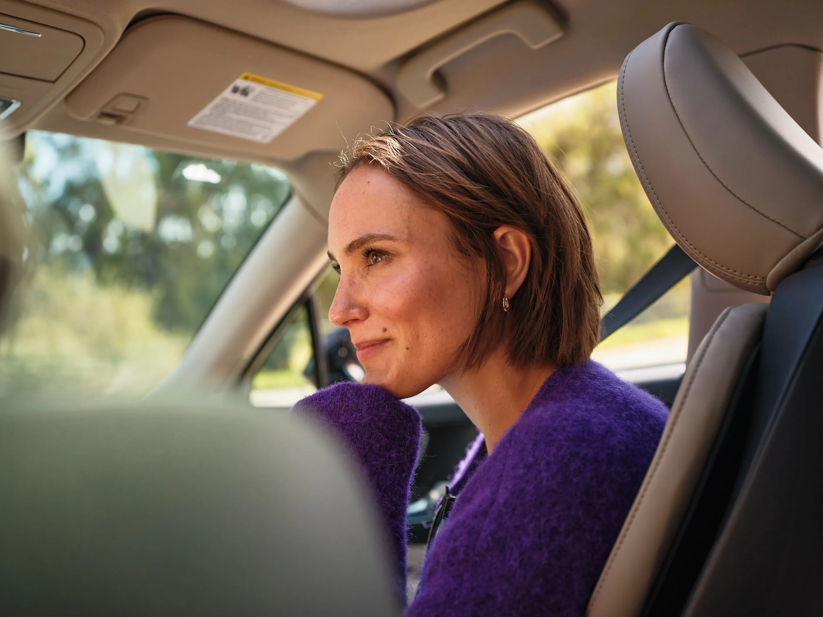 Smiling woman with earrings and purple straps holding her hair back against a clear blue sky.