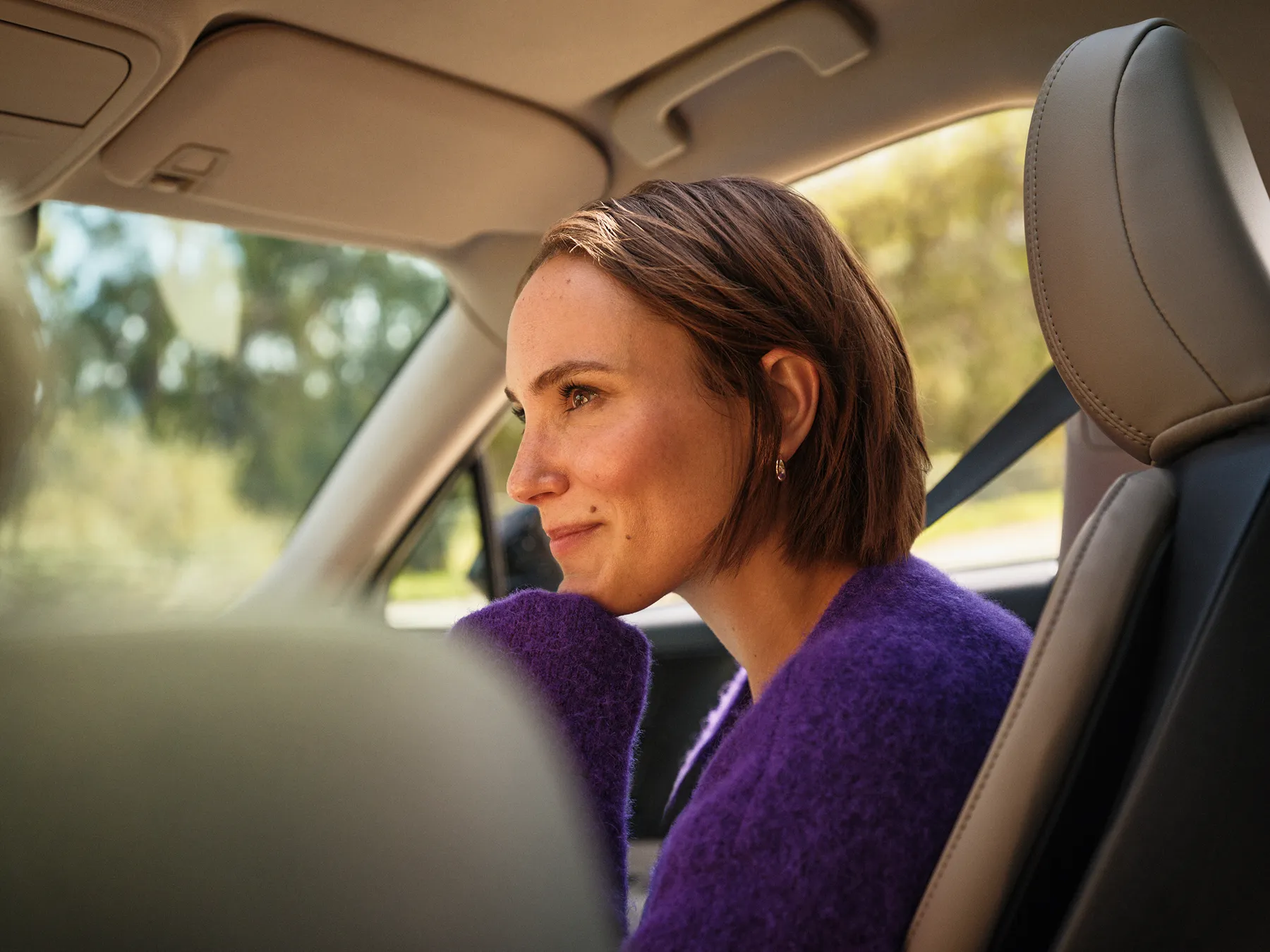 Smiling woman with earrings and purple straps holding her hair back against a clear blue sky.